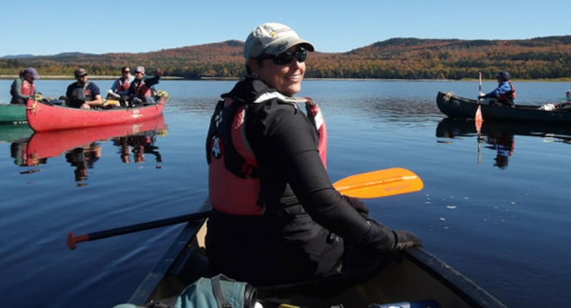 From the back of a canoe, the person in front turns around to smile at the camera. There are additional canoes floating on calm water in the background. 
